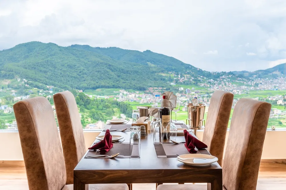 A table laid for four at a window, wine and glassware on dark placemats, terraced fields and a forested hill filling the view