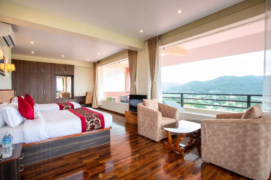 A bed with maroon cushions, two armchairs at a round table by the picture window, and the valley hazy beyond the balcony rail