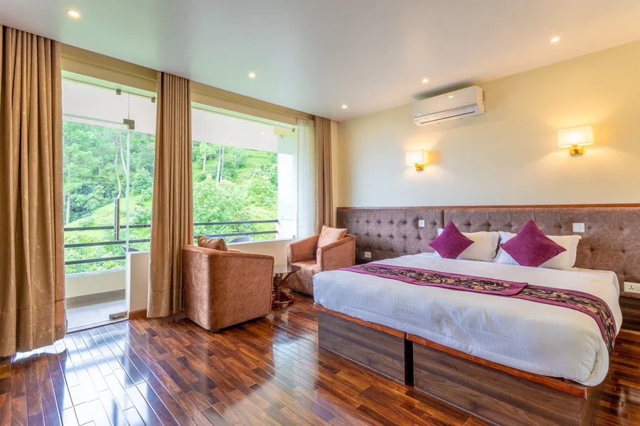 A bed with maroon cushions and runner against a long tufted headboard, polished dark floorboards, and the balcony door open onto greenery