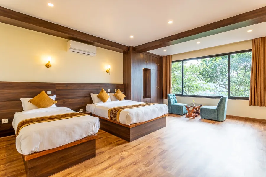 Two beds with gold cushions on a pale timber floor, a sage-green armchair by the window and the trees beyond it