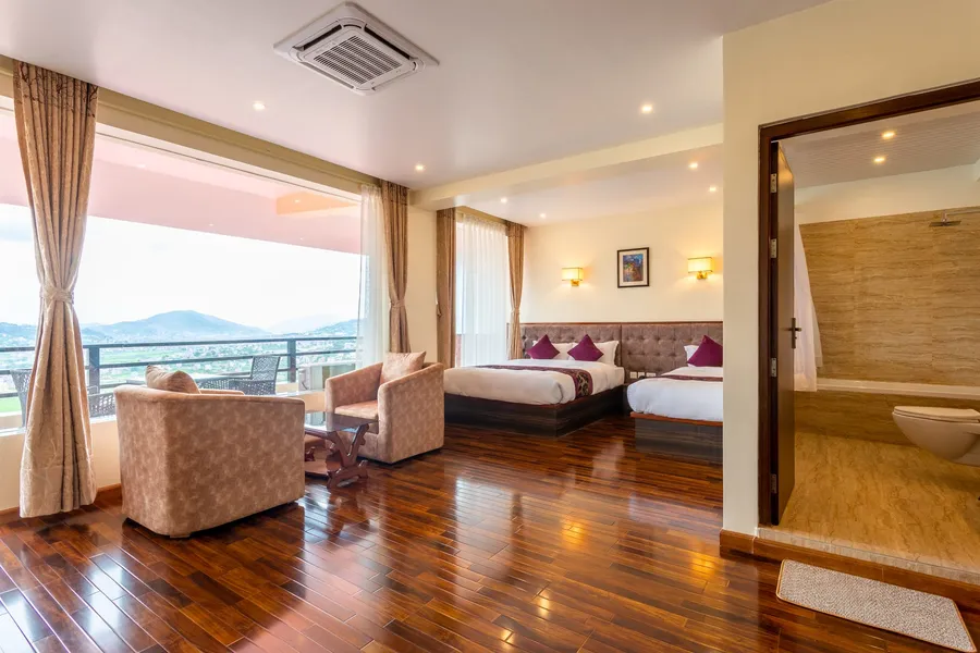 A corner suite glazed on two sides, twin beds with plum cushions against a panelled headboard, armchairs and a carved table at the window wall, the travertine bathroom standing open on the right