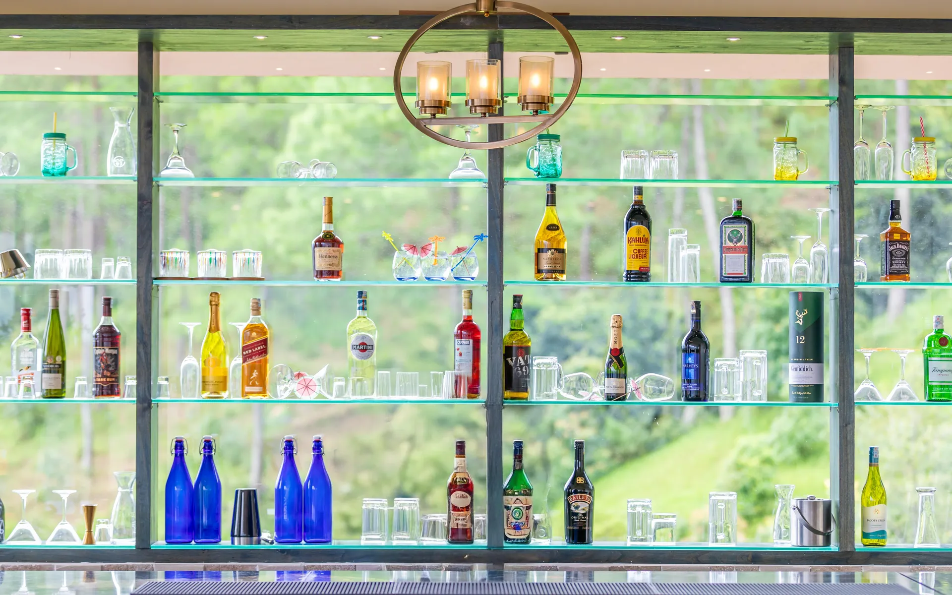 The glass back-bar against the forest windows: lit shelves of bottles and glassware over green shelf edges, cobalt bottles along the counter, the pines beyond