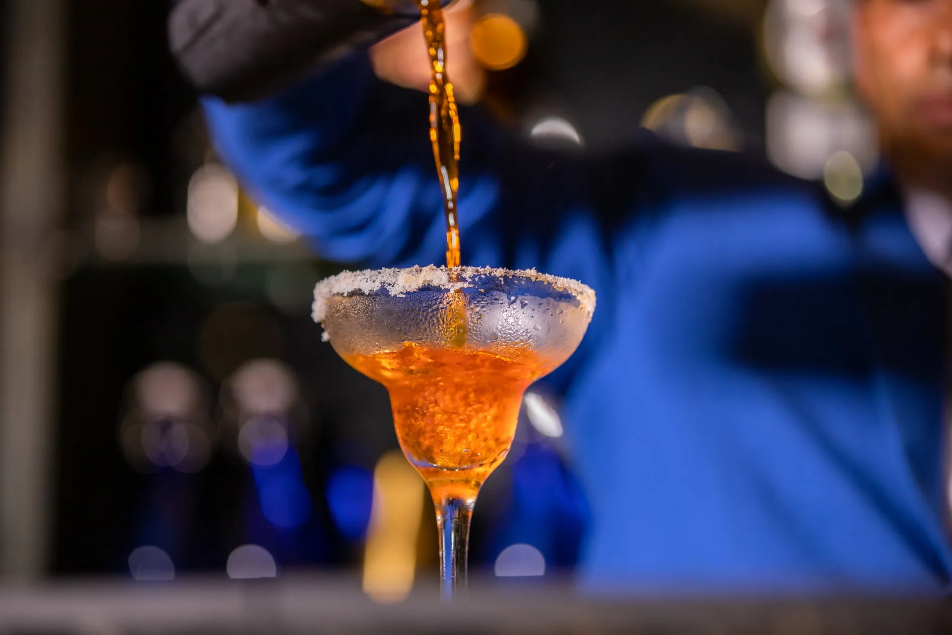 A bartender in a blue jacket pouring a drink into a salt-rimmed cocktail glass, the bar's lights blurred behind him
