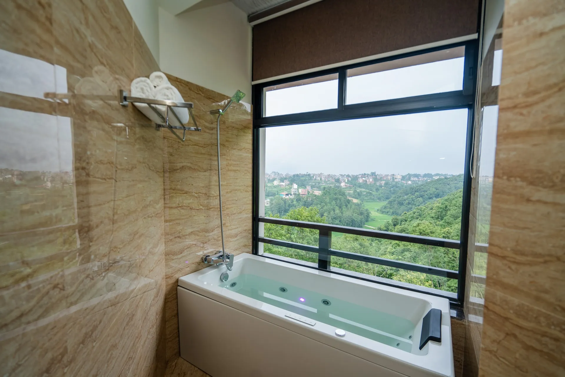 A whirlpool bath set below a picture window in a marble-clad bathroom, folded towels on a rail, the town and wooded hills of the valley beyond the glass