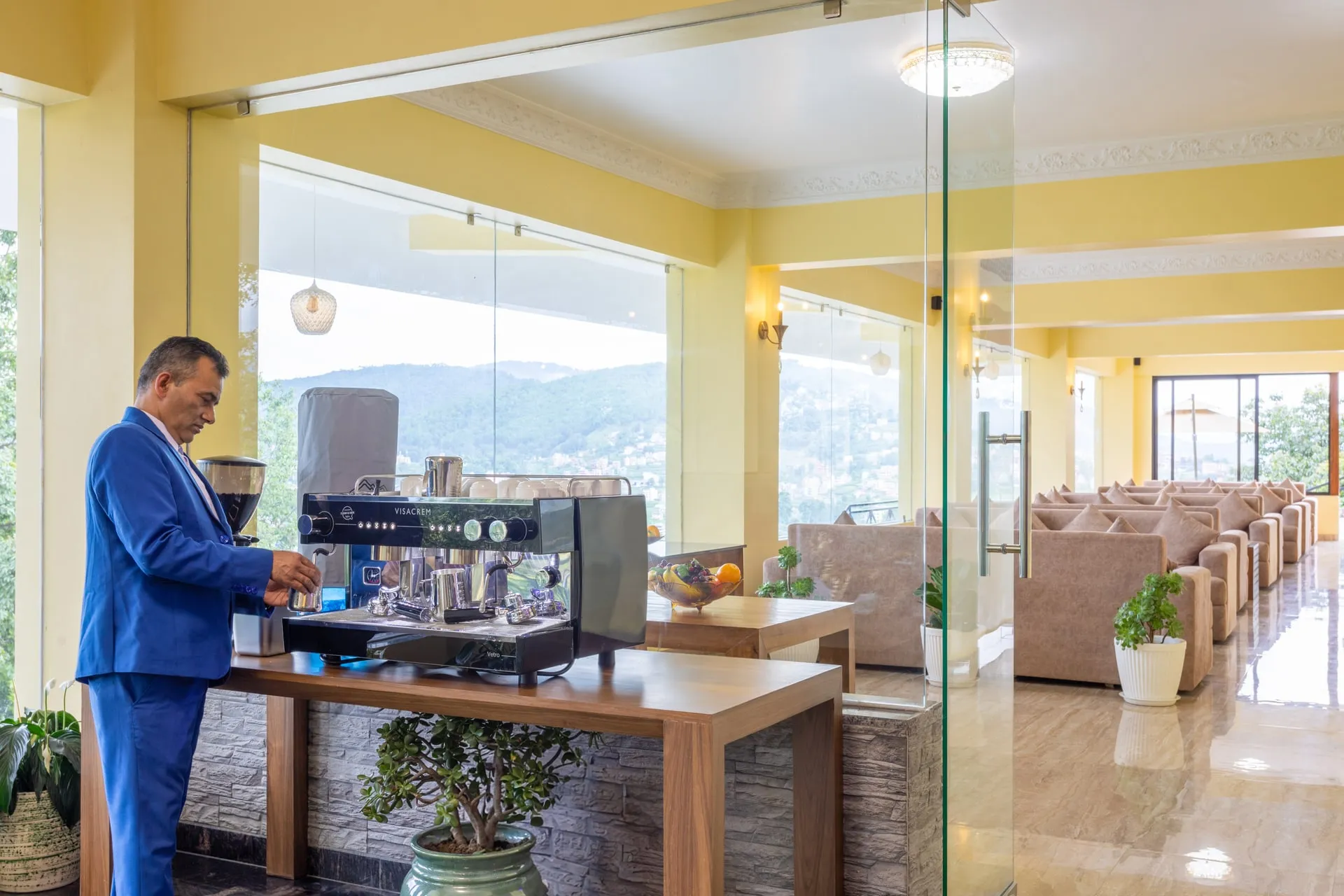 A barista in a blue jacket at the espresso machine on the cafe's wooden counter, the hills through the window wall behind him and the beige sofas of the lounge beyond a glass partition