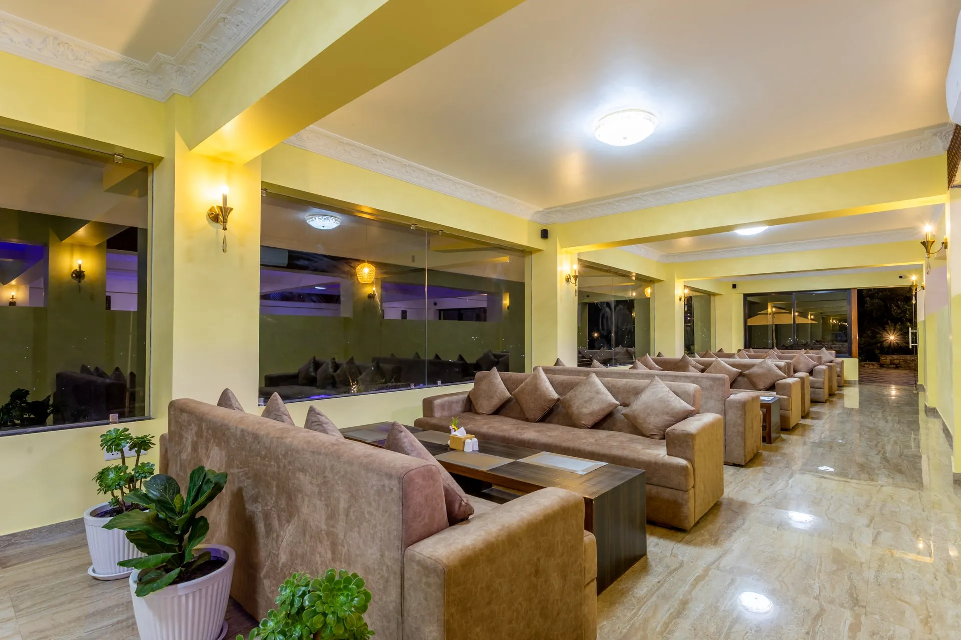 The cafe lounge lit for the evening: rows of beige sofas with cushions along a polished marble floor, yellow walls with wall lamps, potted plants in the foreground and the windows dark