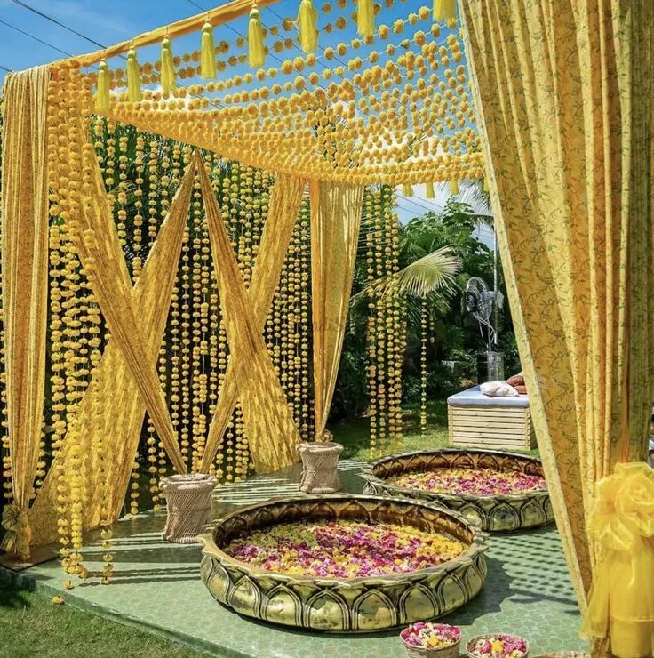 A haldi setting massed in yellow: a canopy of marigold strands and tassels over brass urlis filled with rose and marigold petals, yellow drapes drawn back and garden greenery behind