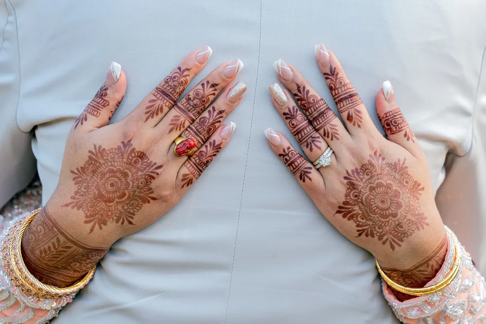 Two hands resting palm-down on pale grey cloth, covered in fresh mehendi from fingertip to wrist — floral medallions on the backs, patterned fingers, gold bangles and rings at the wrists
