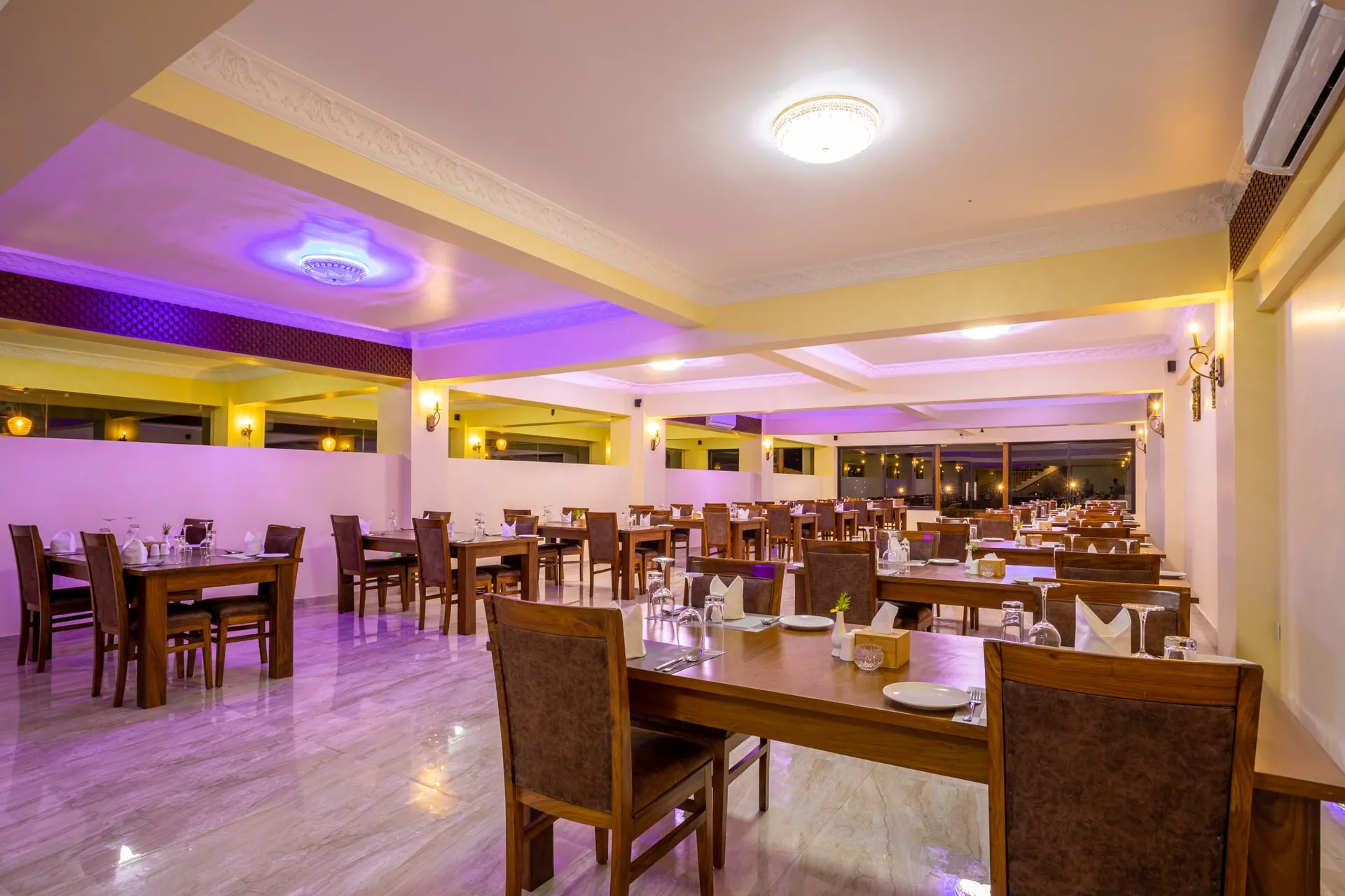 The restaurant laid for service: wooden tables and upholstered chairs set with glasses and folded napkins, the ceiling medallions lit violet and the window wall at the far end