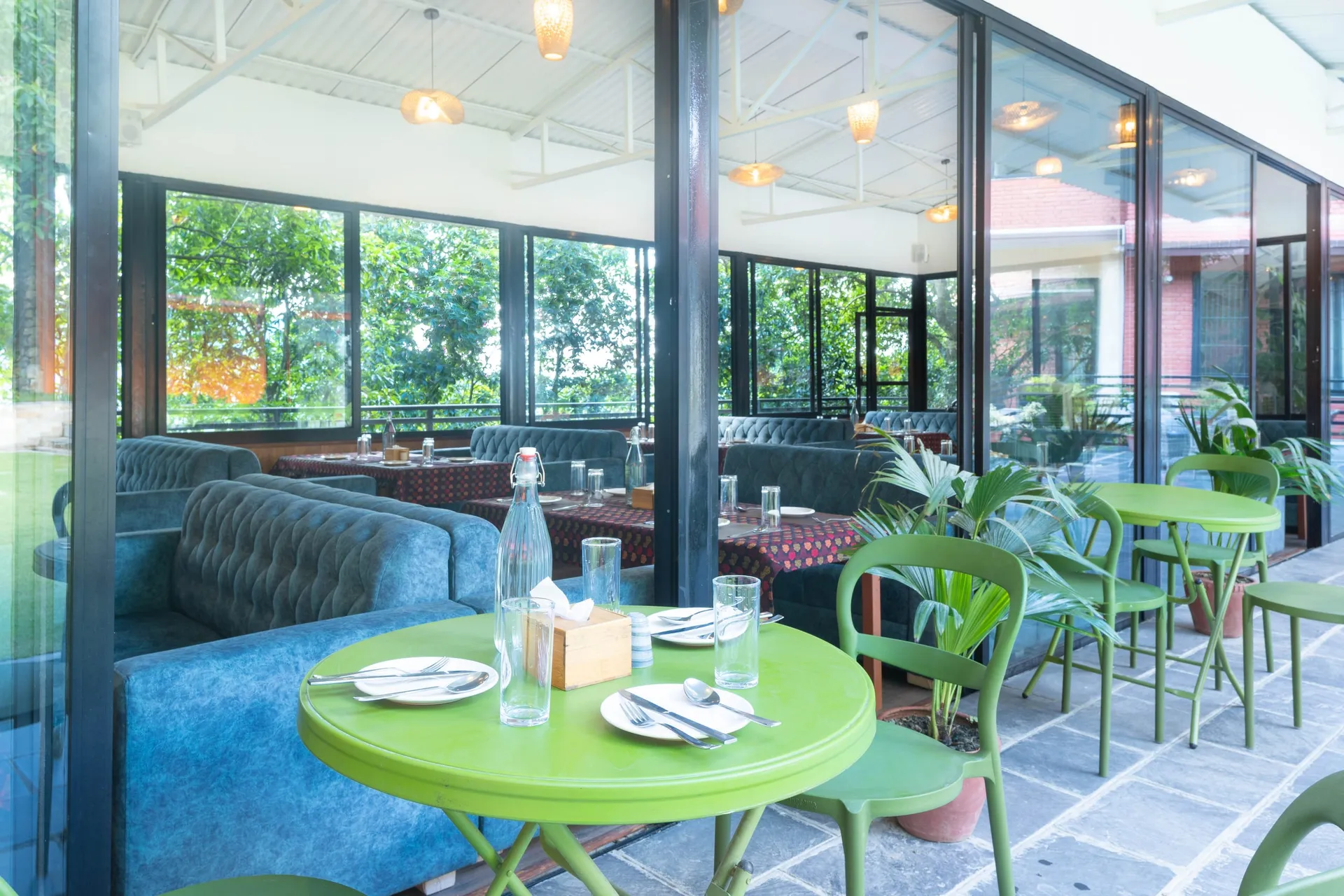 A laid green cafe table and chairs on the paving outside The Nest, the glass wall beside it showing the blue banquettes within and the forest beyond