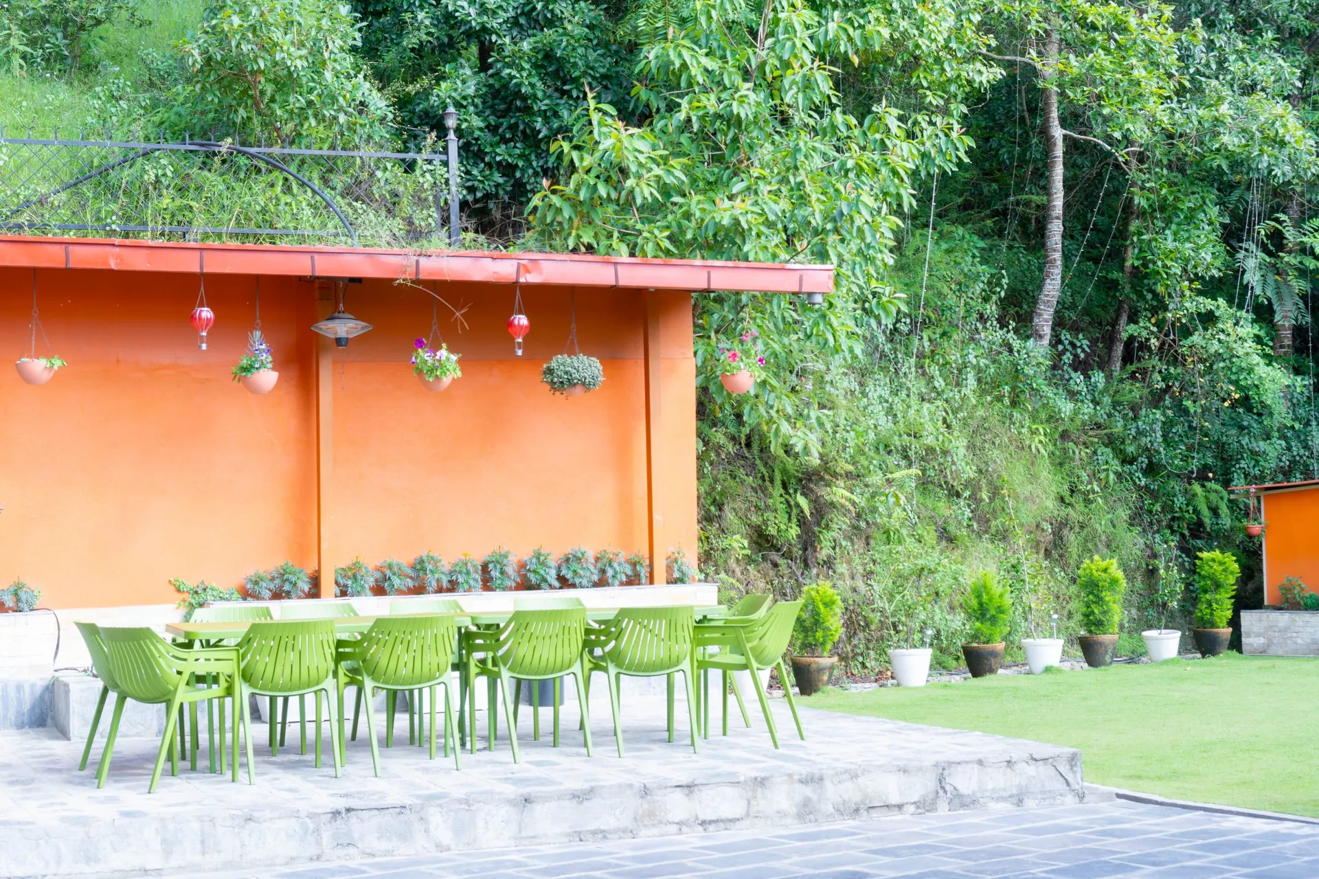 A long table lined with lime-green chairs on the stone terrace below The Nest's orange wall, hanging planters and lanterns above it and forest rising behind