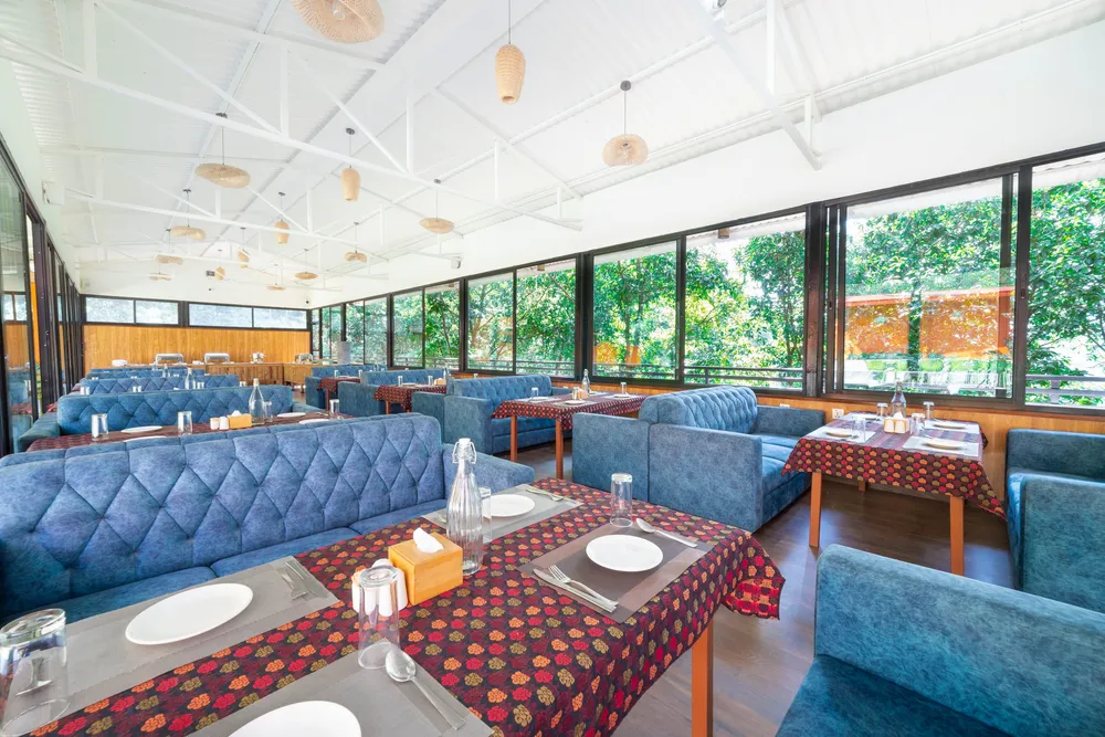 Blue tufted banquettes at tables in red floral cloth under a white pitched roof hung with woven cane lamps, glass walls onto the forest