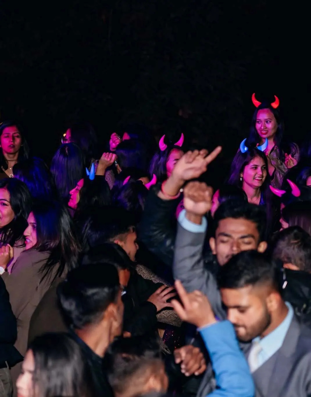 A crowd packed onto an outdoor dance floor at night, hands in the air and glowing horn headbands lit pink and blue against the dark