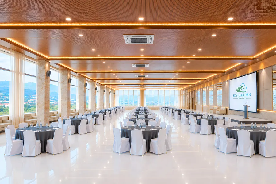 The hall under a slatted timber ceiling with lit coves, set with round tables in charcoal linen and white-covered chairs, water bottles on every table, a screen showing the resort's logo and a lectern on a low stage, window walls onto the valley at the far end