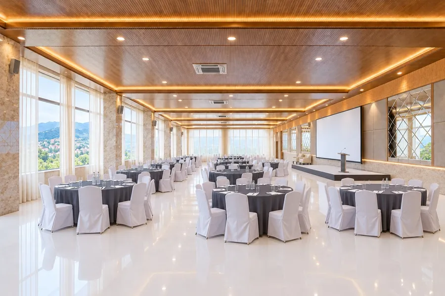 The hall set for a conference under a slatted timber ceiling with lit coves: round tables in charcoal linen with white-covered chairs, water bottles and notepads at every place, a screen and lectern on a low stage, sheer curtains along the window wall onto the valley