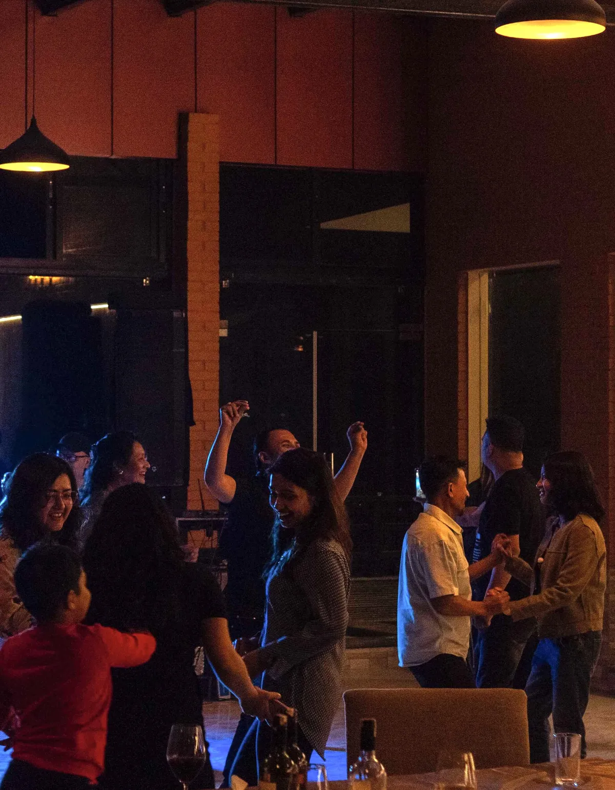 Guests dancing and talking at an evening reception, warm pendant lamps overhead and a brick pillar behind them