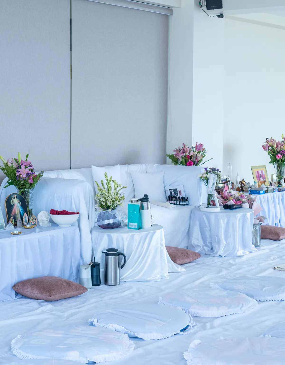 A hall cleared to the floor and dressed in white for a retreat, cushions and mats laid out around draped side tables with flowers, flasks and an altar of small figures and crystals