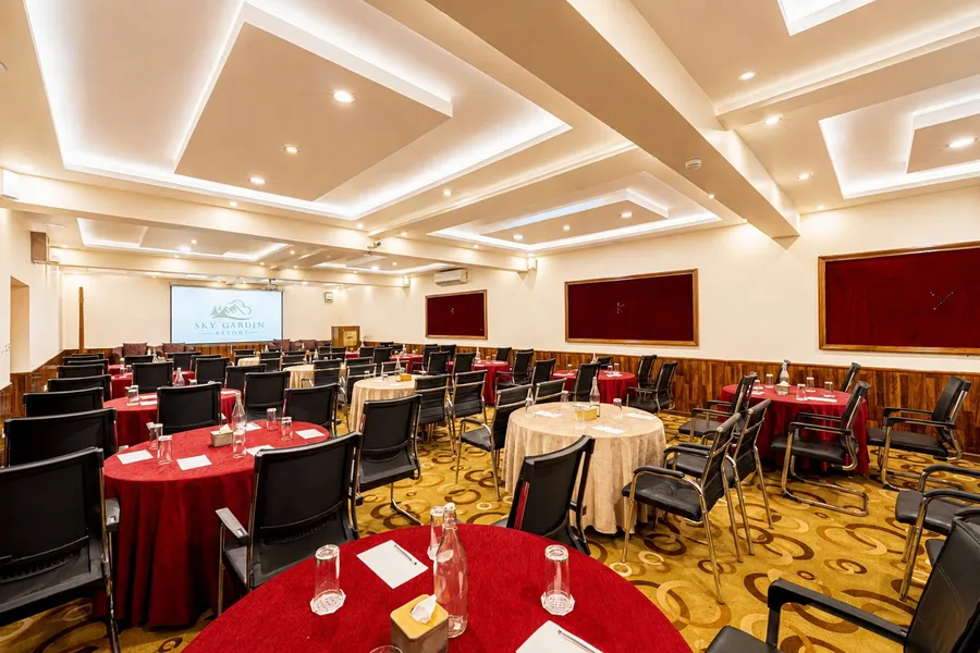 Rosewood laid with round tables in red and gold cloths, black mesh chairs at every table, water carafes and notepads set out, a projection screen and lectern at the far end under a stepped white ceiling