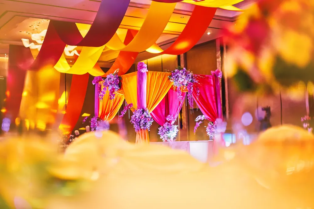 A sangeet dressed in full colour: ribbon streamers in yellow, red and orange swept across the ballroom ceiling, a canopy draped in orange and pink hung with purple orchid clusters, the foreground a warm blur of lights