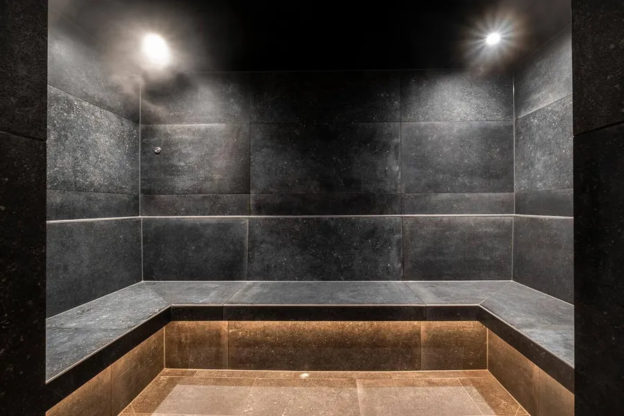 The steam room, dark stone benches wrapping three walls under two ceiling spotlights