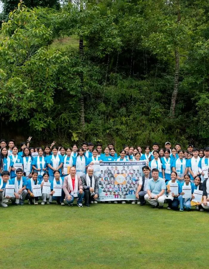 A large group photographed on the resort lawn, rows of guests in matching blue scarves holding certificates around a printed banner, dense forest on the slope behind