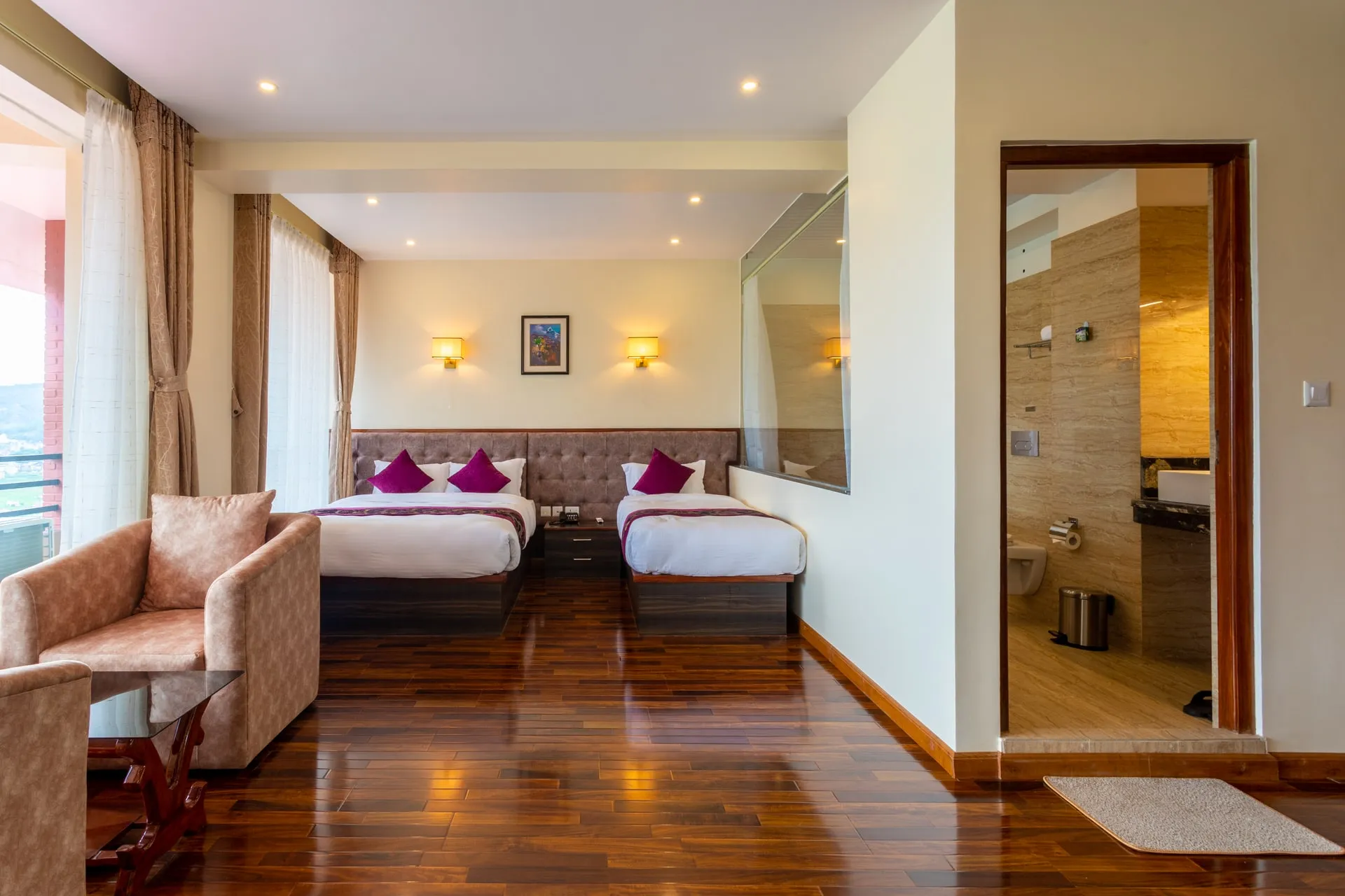 A twin-bedded room: two beds against a tufted headboard with magenta cushions, an armchair by the balcony curtains, a polished wood floor and the marble bathroom through an open door