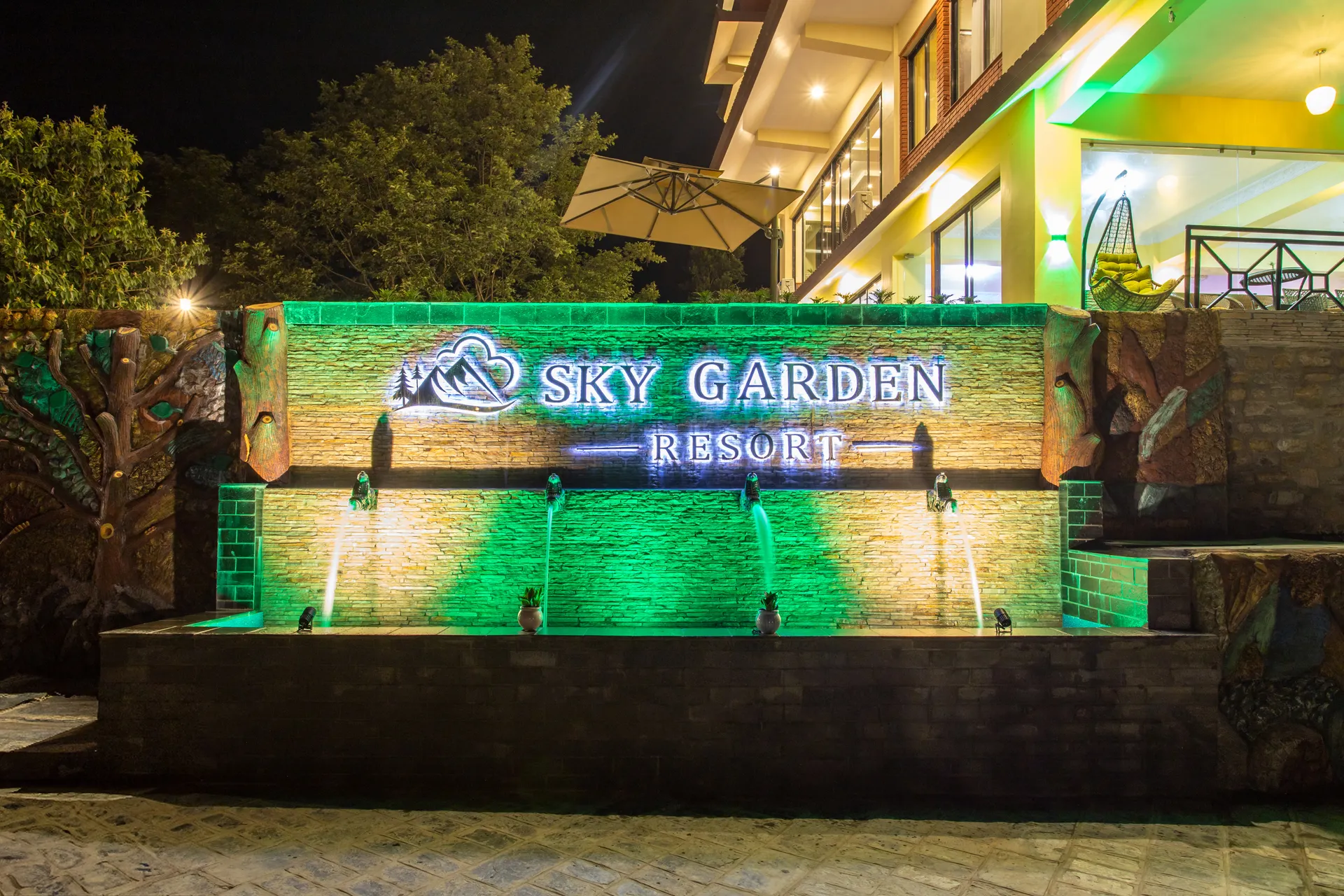The arrival water wall lit after dark: four jets arcing into a basin below the stone Sky Garden Resort signage, the wall washed green and gold, carved tree reliefs at either end and the lit terrace of the resort behind