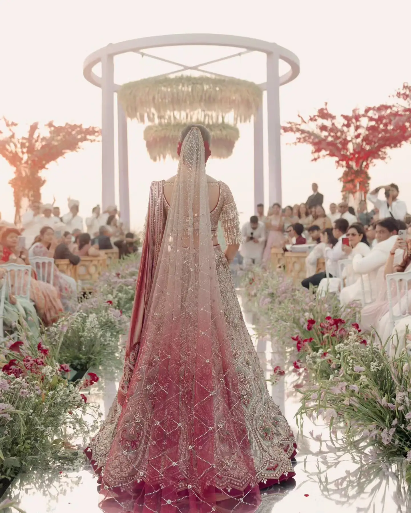 A bride in a deep-rose embroidered lehenga and a long veil walking a mirrored aisle towards a white circular mandap hung with two tiers of blossom, banks of flowers down both sides of the aisle and seated guests beyond
