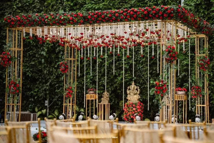 A mandap of slim gold posts against a wall of greenery, its canopy massed with red roses and hung with strands of white blossom, a garlanded idol at the centre and rows of gold chairs in front