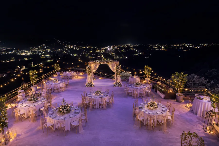 The rooftop terrace dressed for an evening reception: round tables under white cloths with gold chiavari chairs, a draped floral arch at the parapet, festoon lights overhead and candles along the edge, the lights of the valley spread out below