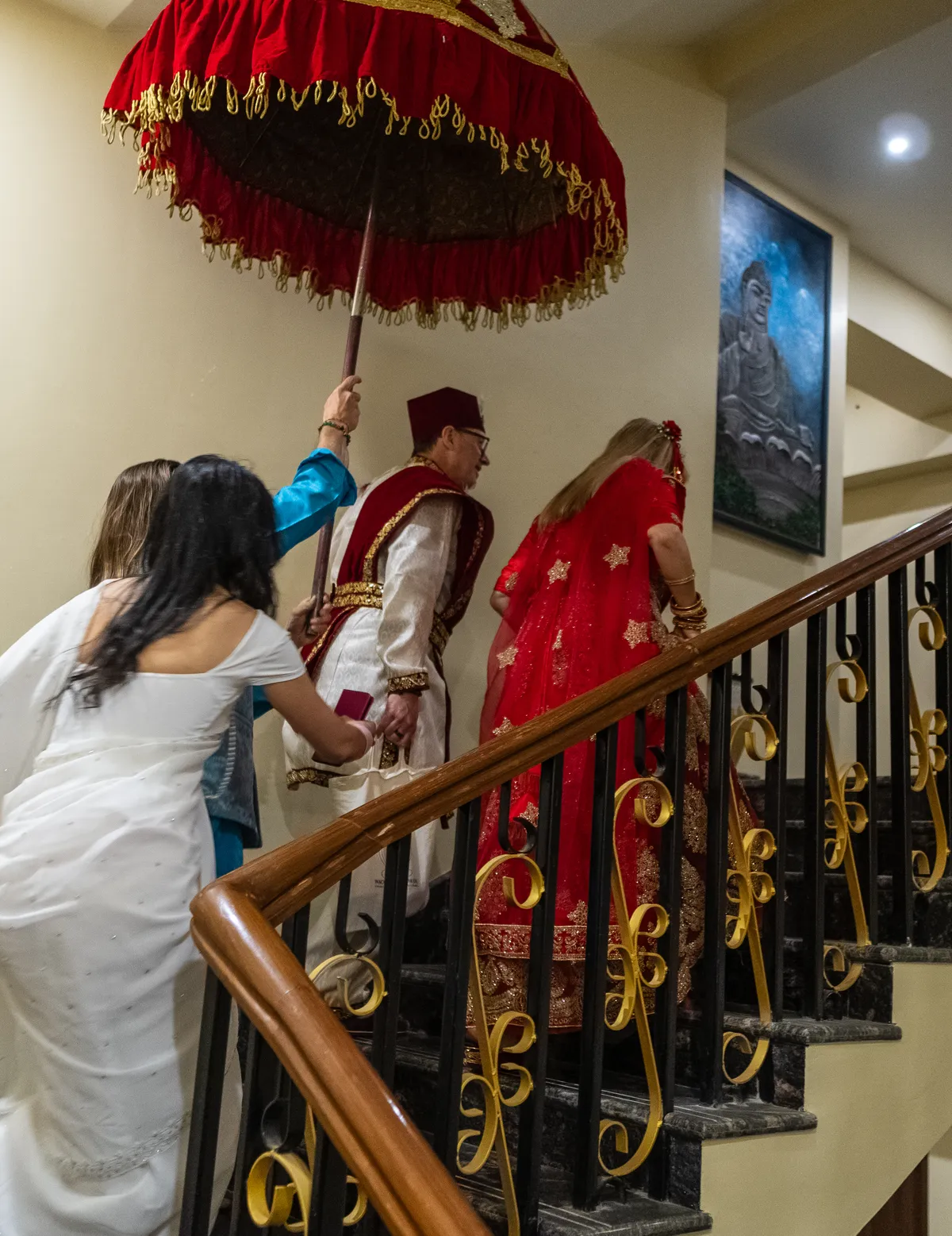 A wedding party climbing an interior staircase beneath a red and gold ceremonial parasol, the bride in a red and gold sari and the groom in a topi and traditional dress