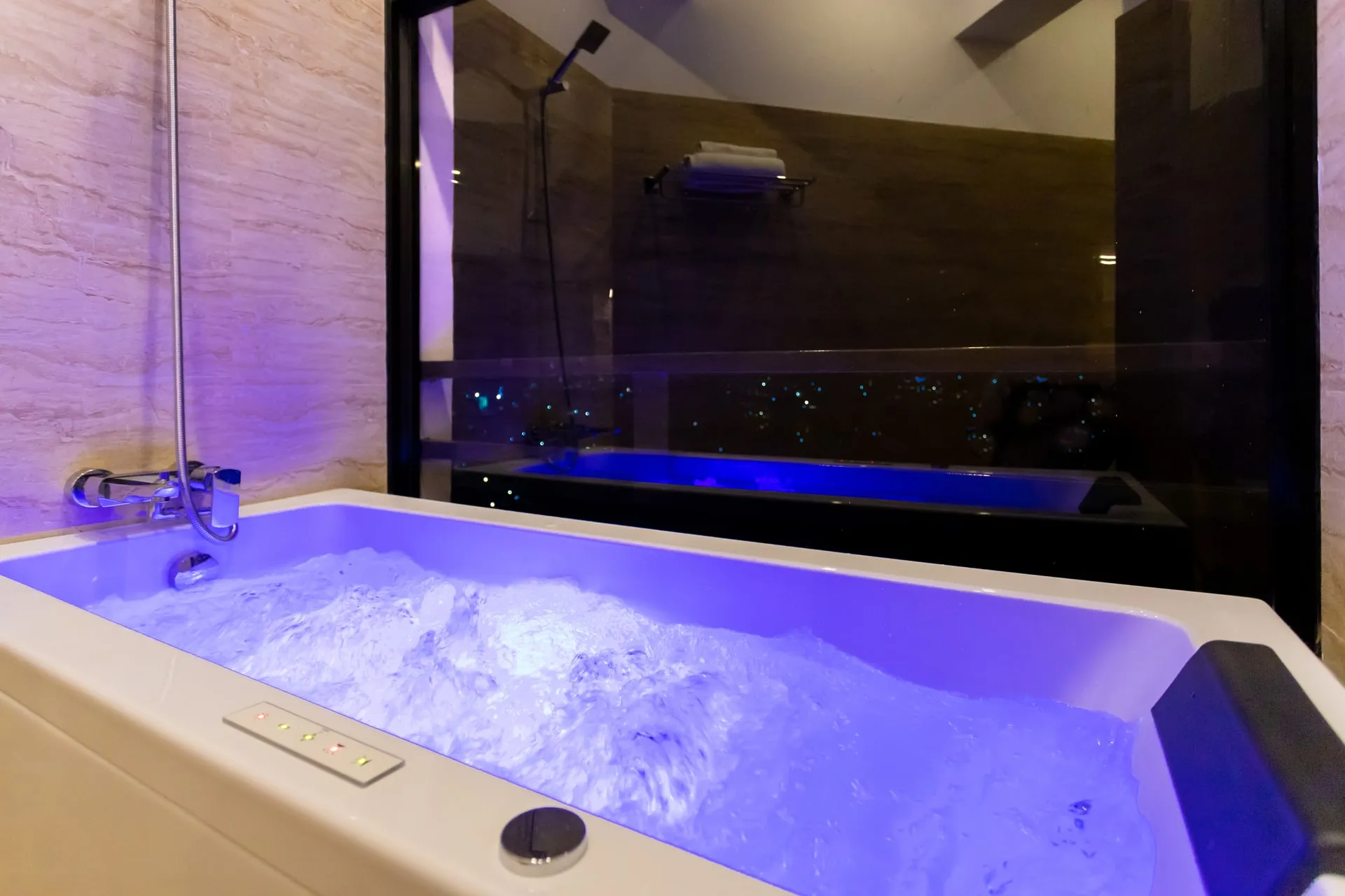 A whirlpool bath running, its water lit blue, a shower fitting on the marble wall beside it and a dark glass window behind